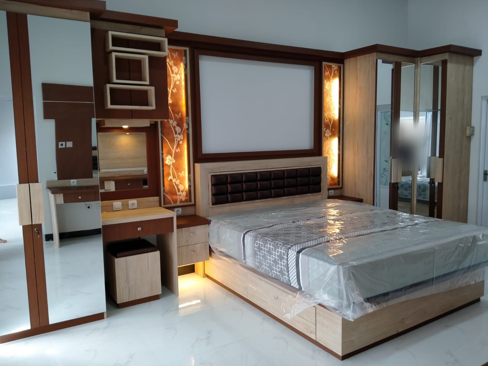 Kamar tidur dengan ranjang berlaci, headboard berlapis kain, panel dinding berlampu, meja rias, dan lemari berpintu cermin.