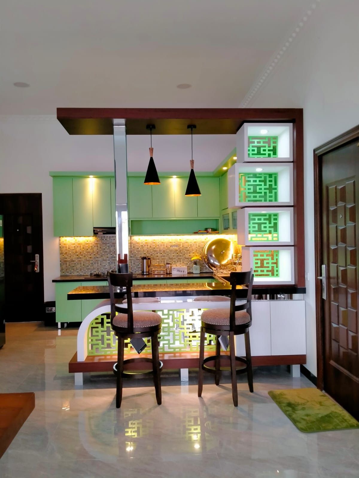 Dapur dengan kabinet hijau muda, meja bar berpanel berlubang, dan rak hias bersusun yang berfungsi sebagai pembatas ruang.