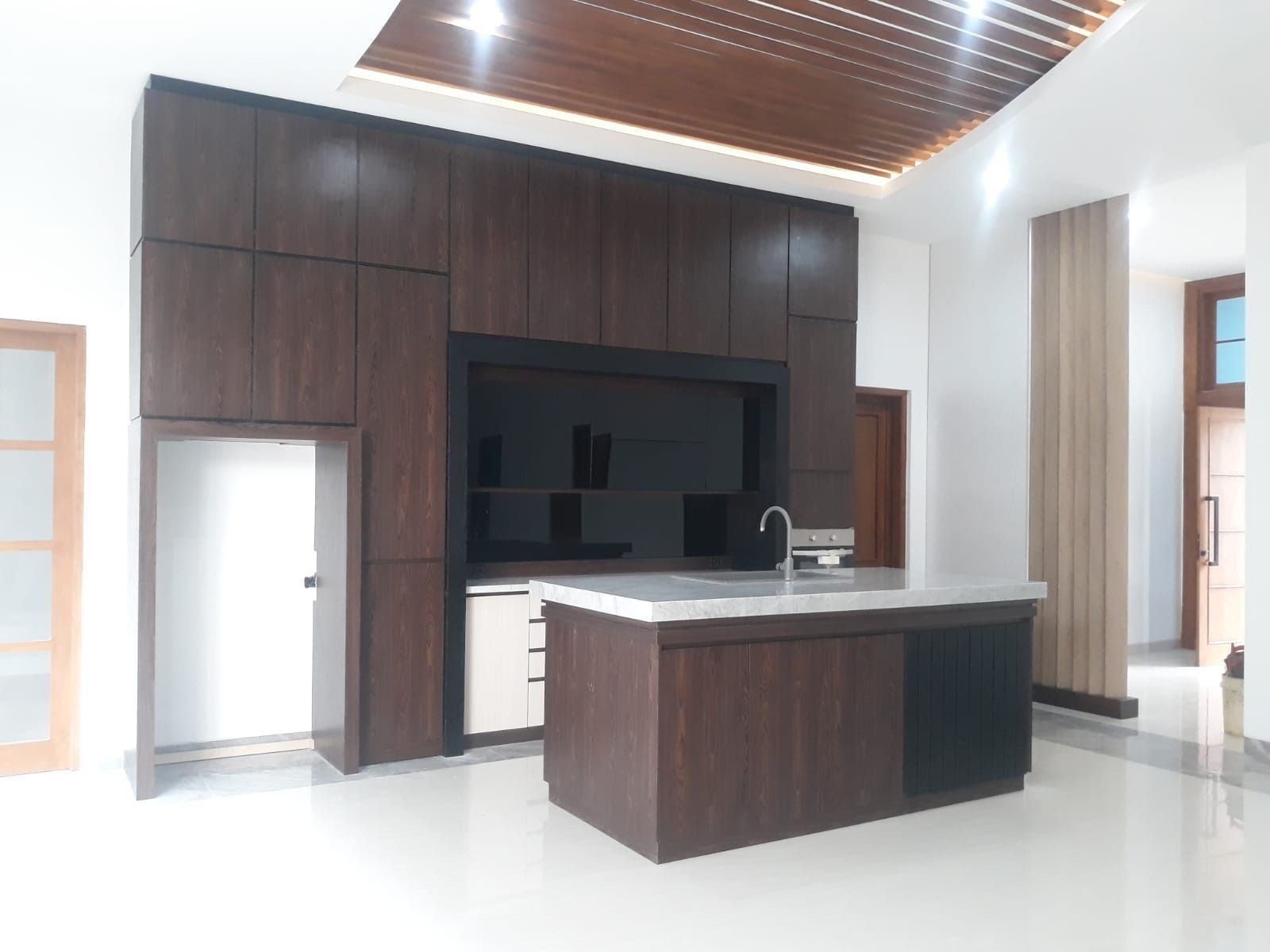 Dapur dengan kabinet kayu gelap setinggi plafon dan island berlapis meja batu putih di rumah yang belum berisi perabot.