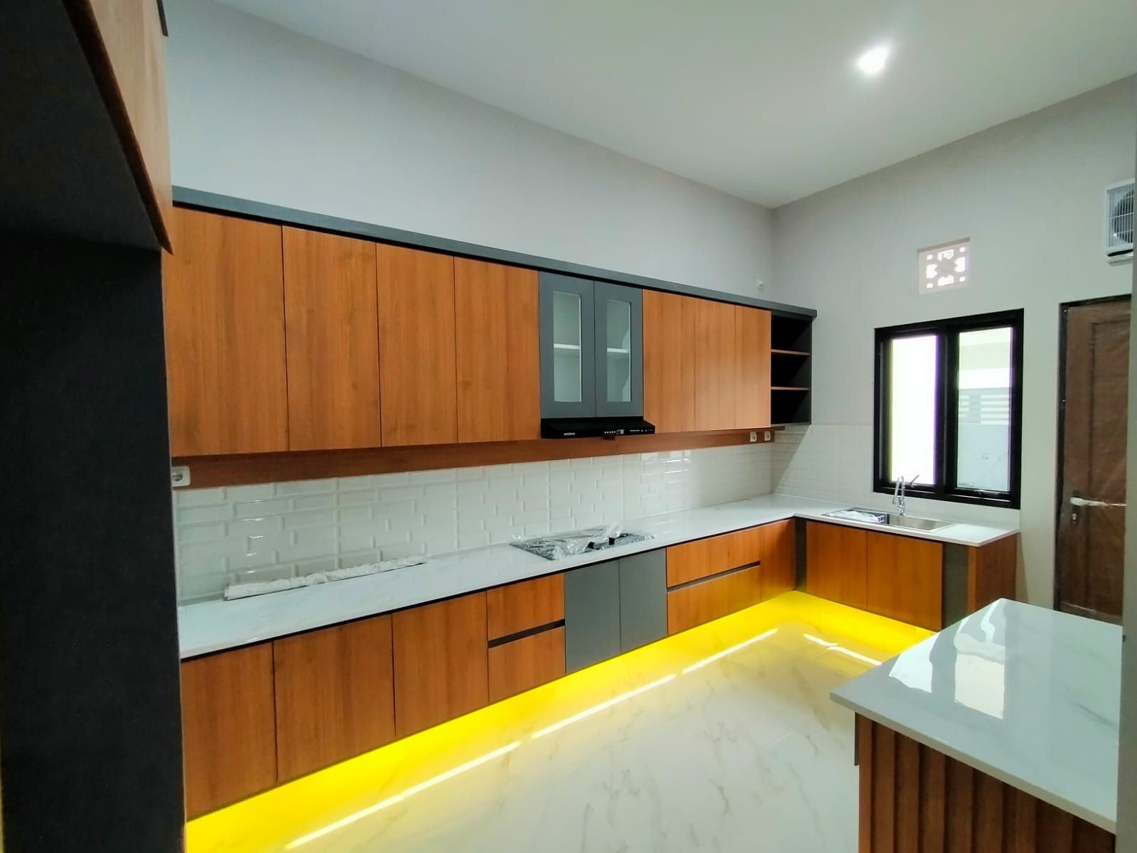 Dapur panjang dengan kabinet bermotif kayu, pintu kaca di atas kompor, dan lampu tersembunyi di bawah kabinet bawah.