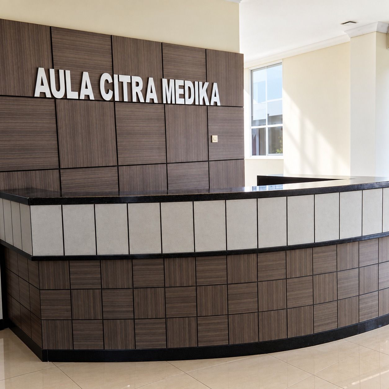 Lobi Aula Citra Medika dengan meja resepsionis melengkung, pita panel terang, dan dinding latar berpanel kotak motif kayu gelap.