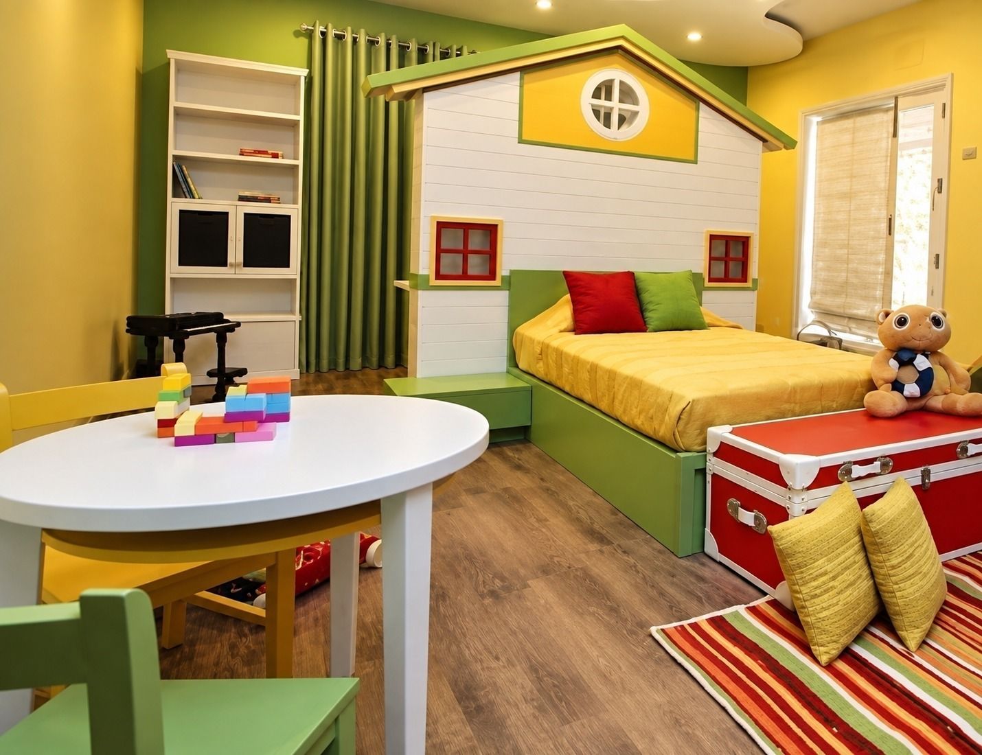 Kamar anak kuning dan hijau dengan ranjang berbentuk rumah beratap, rak tinggi, dan meja bundar.